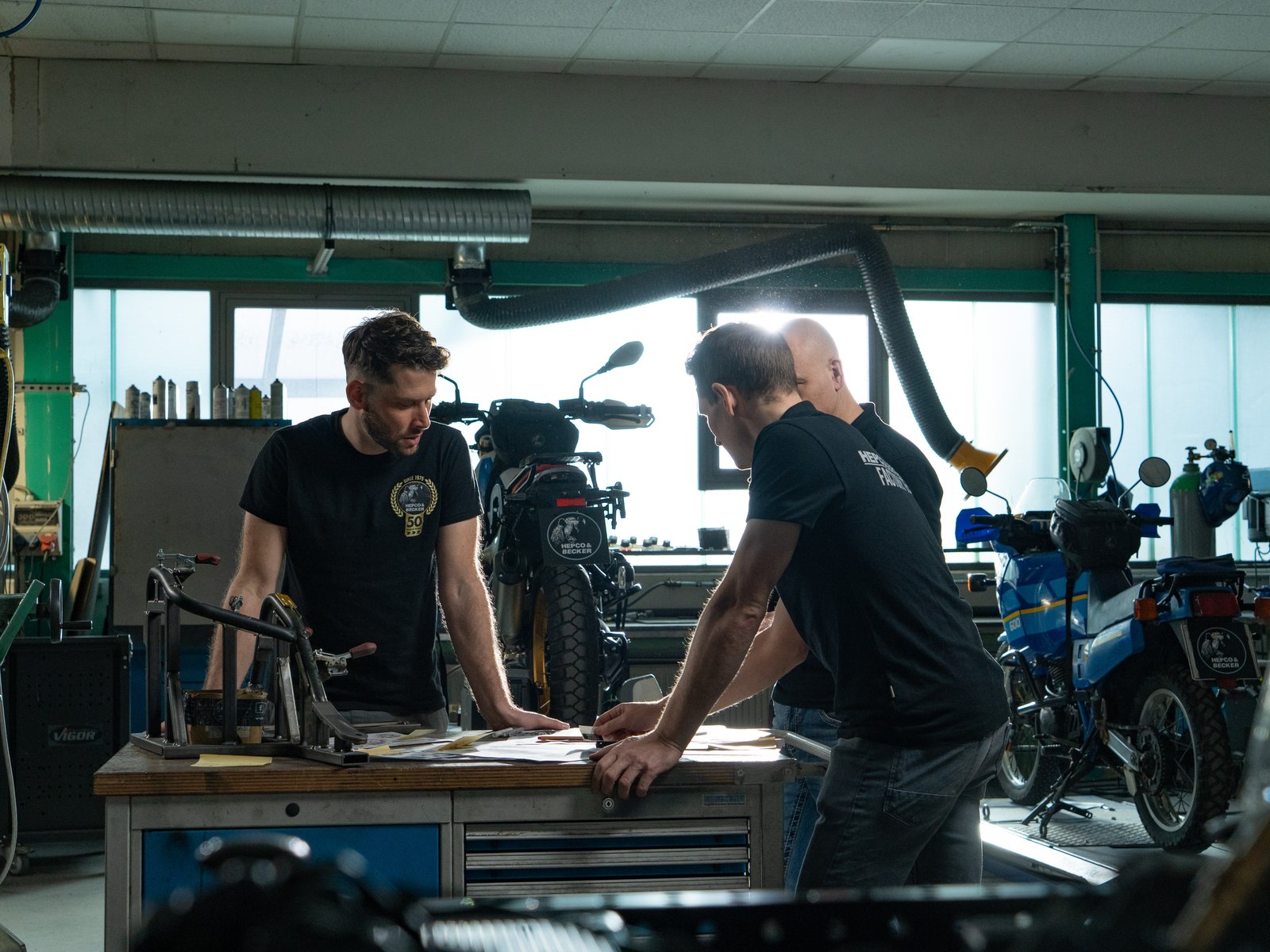 Dwaj konstruktorzy Hepco & Becker pochyleni nad prototypem stelaża na stole warsztatowym, w tle motocykle na podnośnikach w dziale rozwoju w Pirmasens